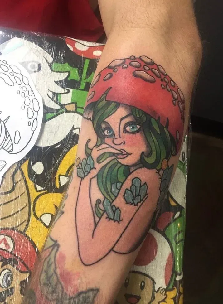Tattoo of a woman with green hair and a red mushroom cap, arms crossed, on a forearm.
