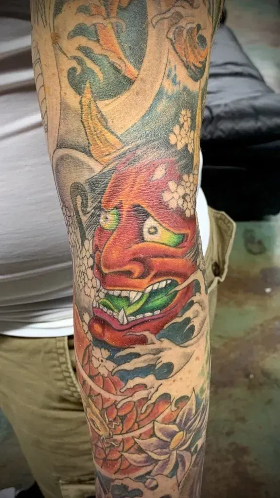 Tattoo of a red demon mask with green eyes and tongue, surrounded by floral and wave designs on an arm.