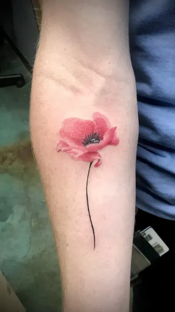 A tattoo of a vibrant red poppy on a person's forearm. The flower has a black center and a thin, curved stem.