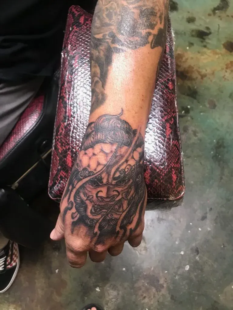 Close-up of a hand with an intricate, dark tattoo covering the hand and wrist. The tattoo design is detailed and flowing.