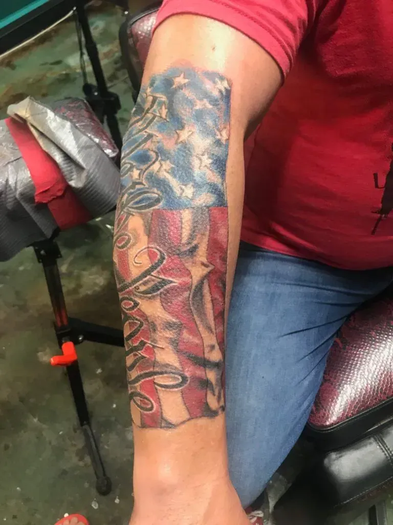 Tattoo of an American flag on a person's forearm, with cursive script; person wearing a red shirt and jeans.