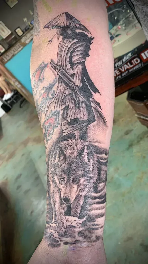 Tattoo on forearm: Wolf below building with a figure in a hat. Black and grey.