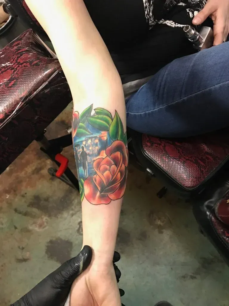 Arm with a colorful tattoo of a rose and other foliage, being examined by a person wearing a black glove.