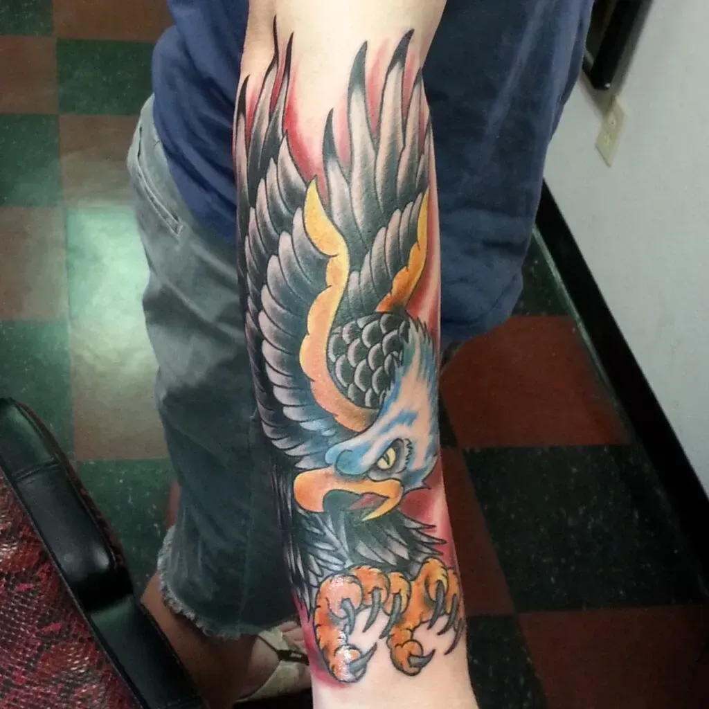 Tattoo of an eagle with outstretched wings on a forearm, with black, gray, orange, and blue colors.