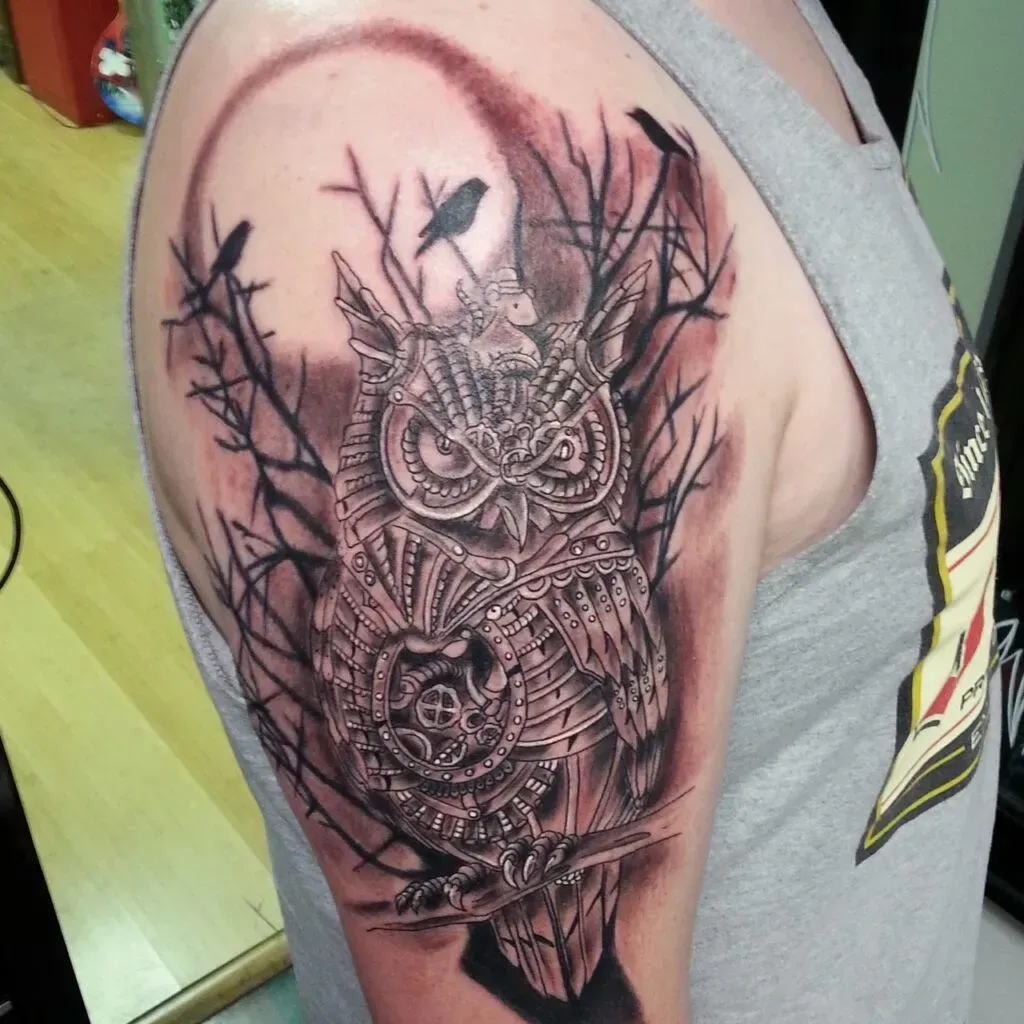 Tattoo of a mechanical owl on a person's upper arm, surrounded by branches and birds, against a crescent moon.