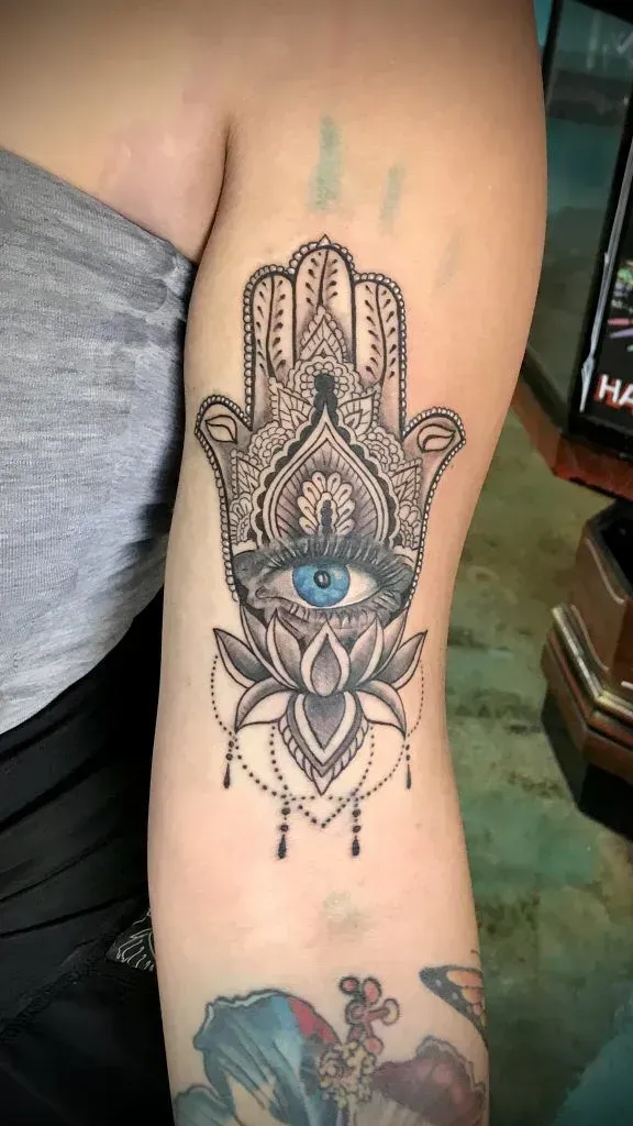 Hamsa hand tattoo with blue eye, lotus flower, and intricate black detailing on a person's upper arm.
