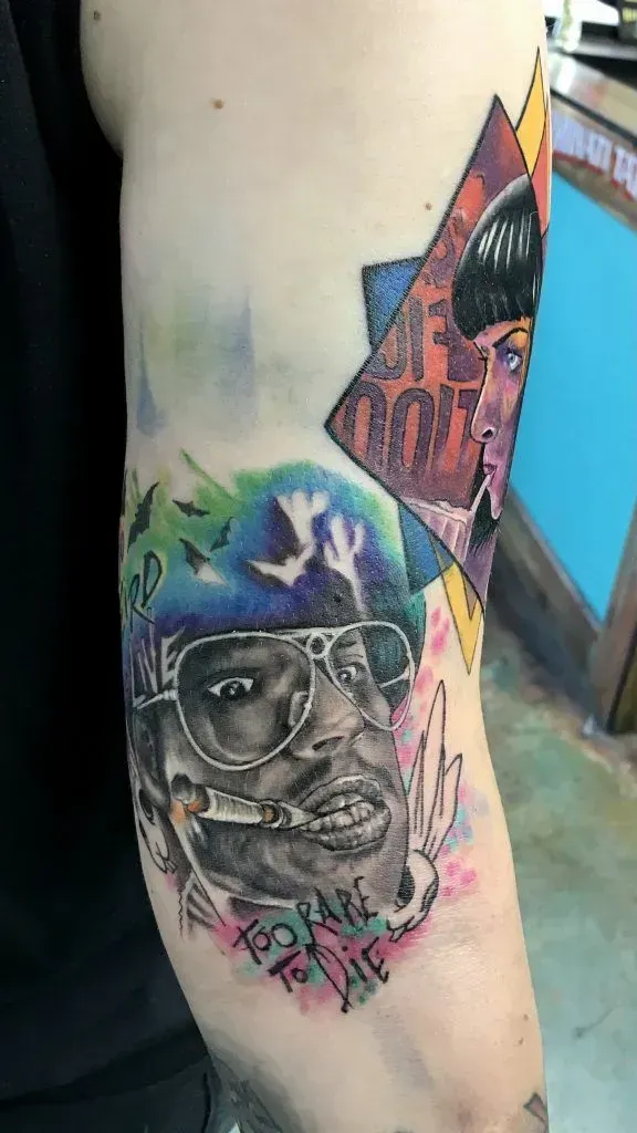 Tattoo on arm, featuring portrait with sunglasses, cigarette, and music poster, with colorful watercolor accents.