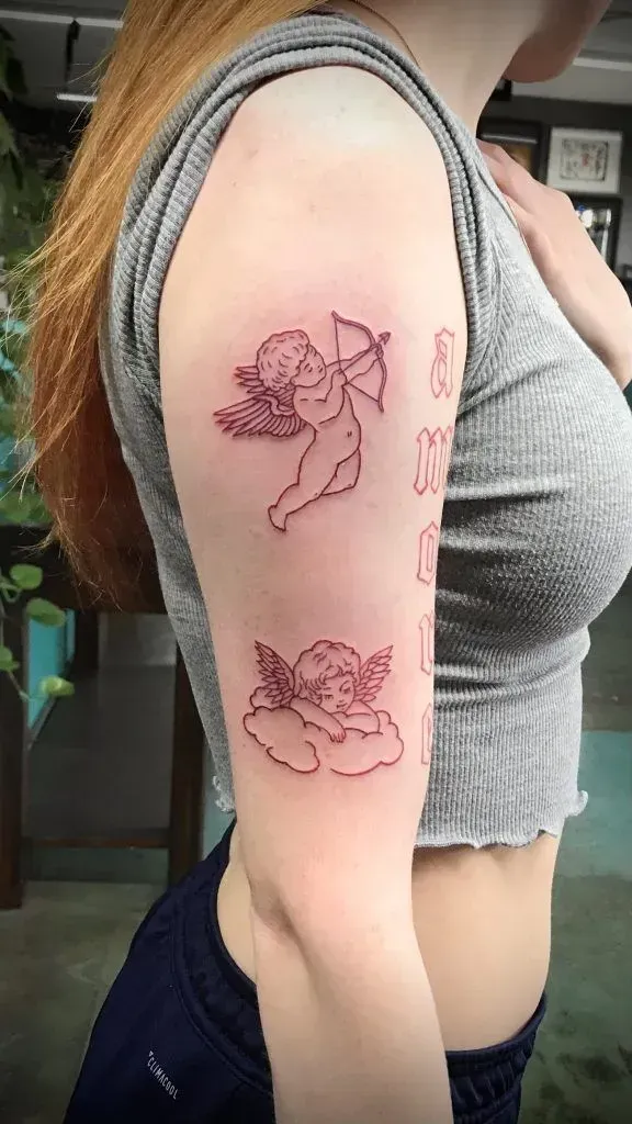 Arm with two outlined cherub tattoos; one with bow, one on cloud. Person wears gray top.