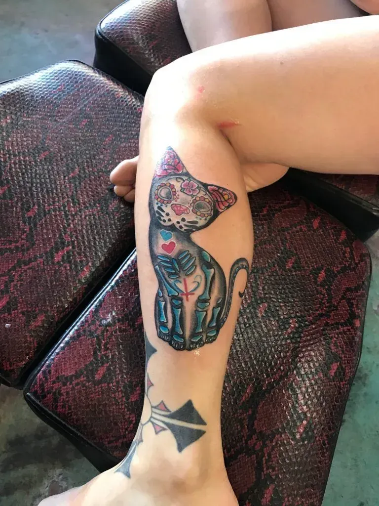 Calf tattoo: cat in Dia de los Muertos style with colorful floral headpiece, skeleton, and sitting on a textured surface.