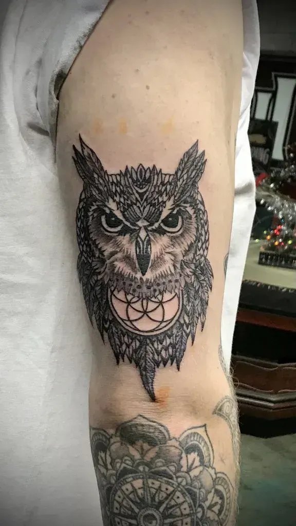 Tattoo of a detailed owl head with geometric design on the upper arm. The owl has an open mouth with a symbol.