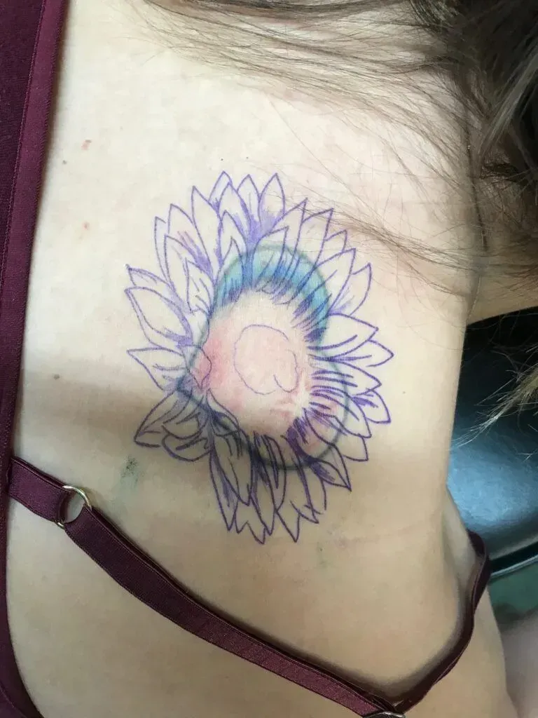 Outline of a sunflower tattoo on a person's upper chest, with blue and pink shading.