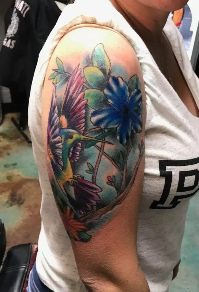 Tattoo of a hummingbird and blue flower on an arm, vivid colors: green, purple, and blue.