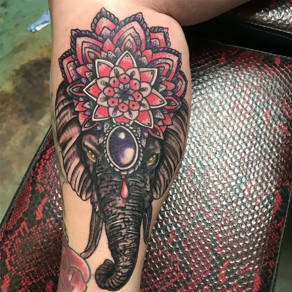 Elephant tattoo with a mandala headpiece, purple jewel, and red accents on a leg.