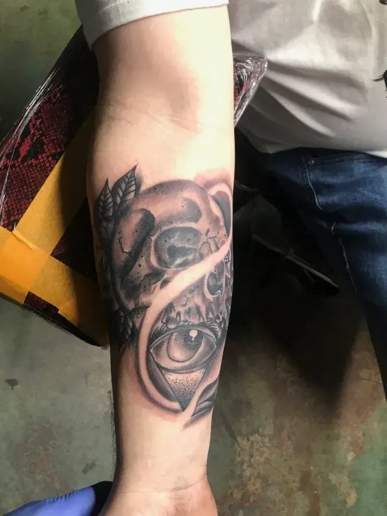 Forearm tattoo: Skull with an eye, shaded in black and grey, surrounded by leaves.