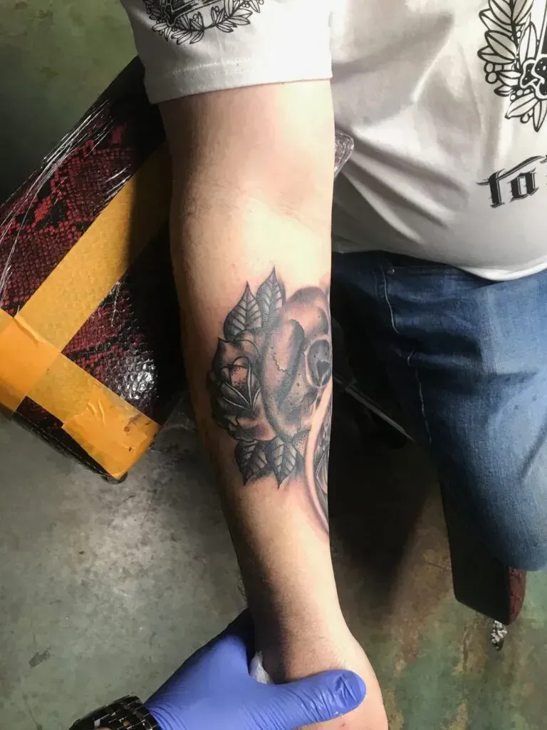 Forearm tattoo of a skull and rose. Person's arm is held by a gloved hand.