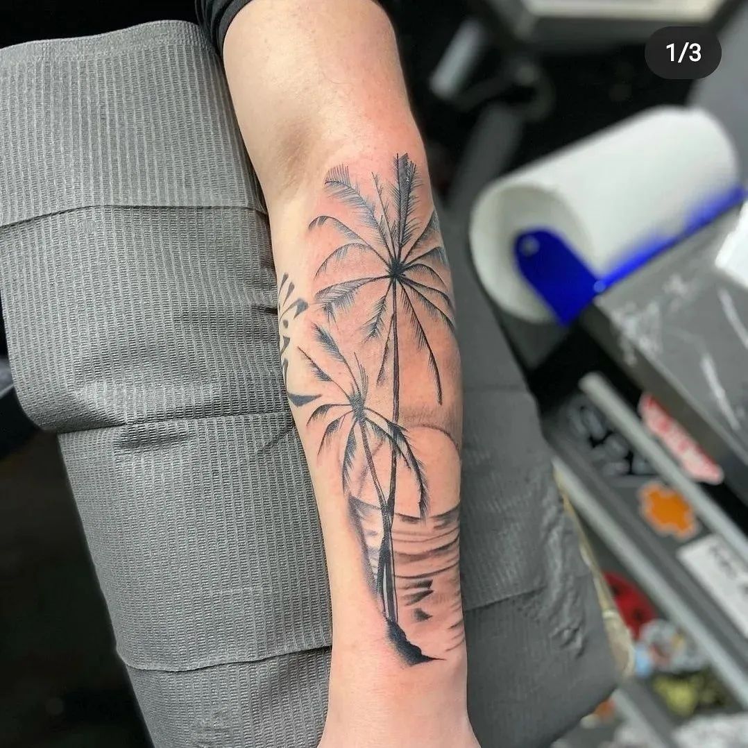 Palm tree tattoo on forearm, black ink, against a sunset, on grey textured background.