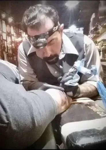 Tattoo artist with headlamp, working on a client's arm. Dark setting, wearing vest, focused expression.