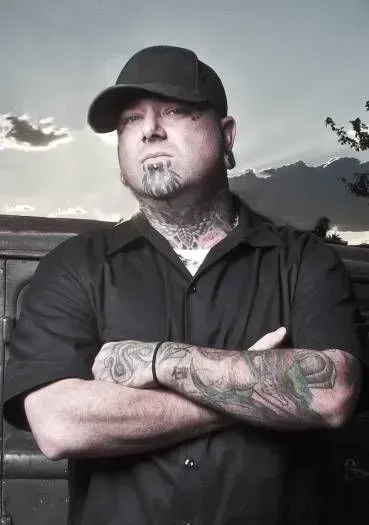 Man with tattoos, wearing a black cap and shirt, arms crossed, standing outdoors.