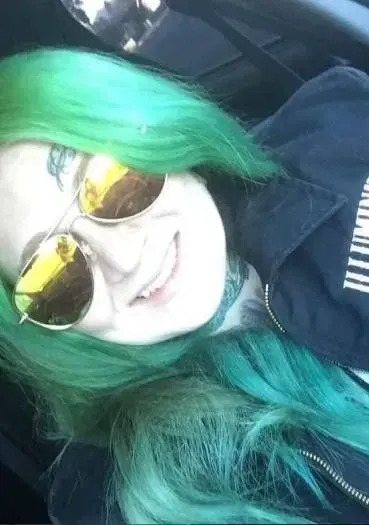 Person with green hair and yellow sunglasses smiles; close-up photo.