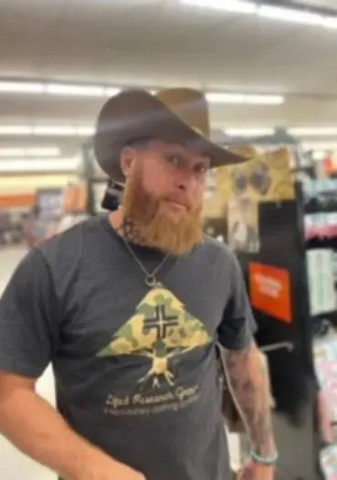 Man in cowboy hat and beard wearing a t-shirt with a tree design, inside a store.