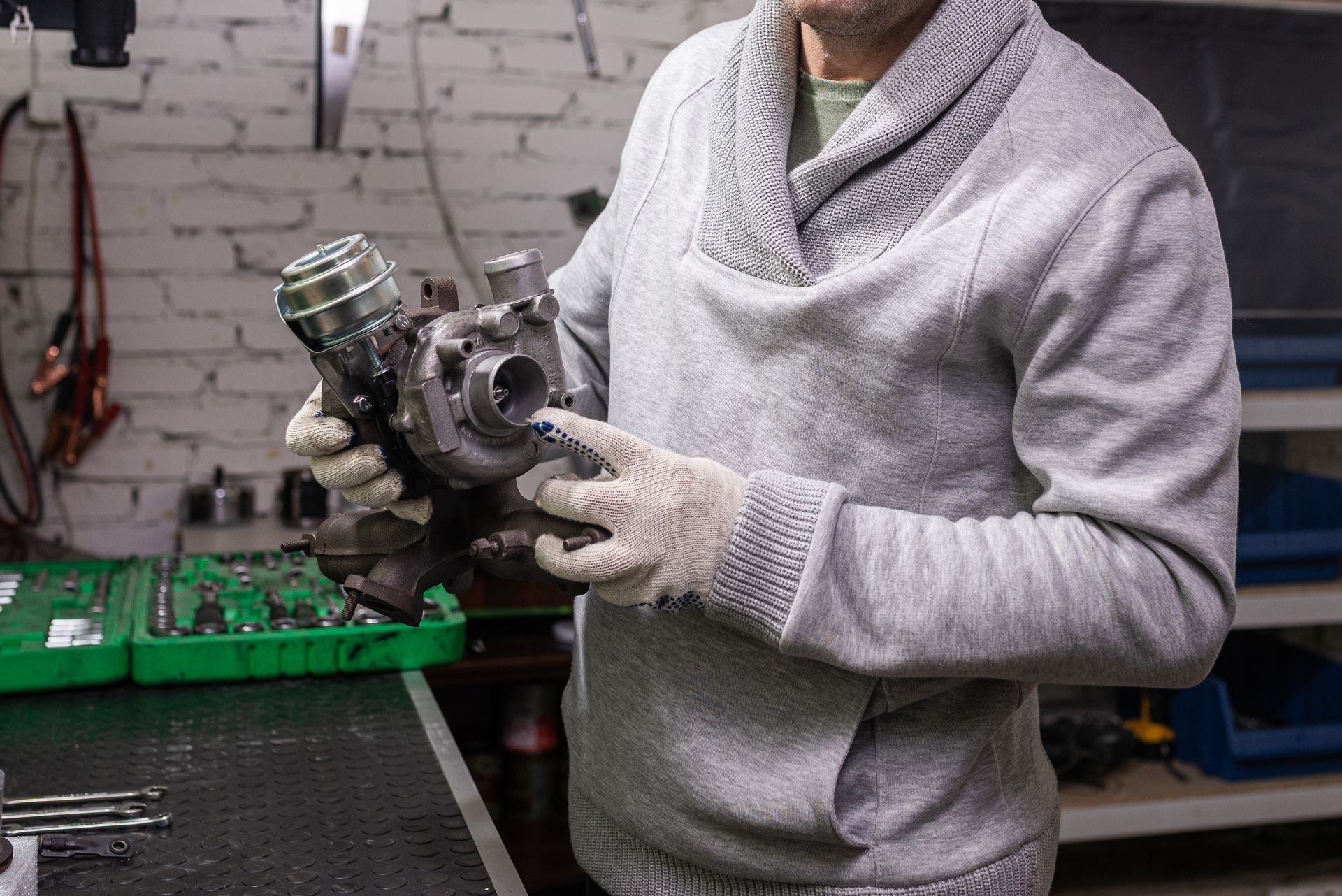 A person in a gray sweater and work gloves inspects a metal turbocharger in a workshop.