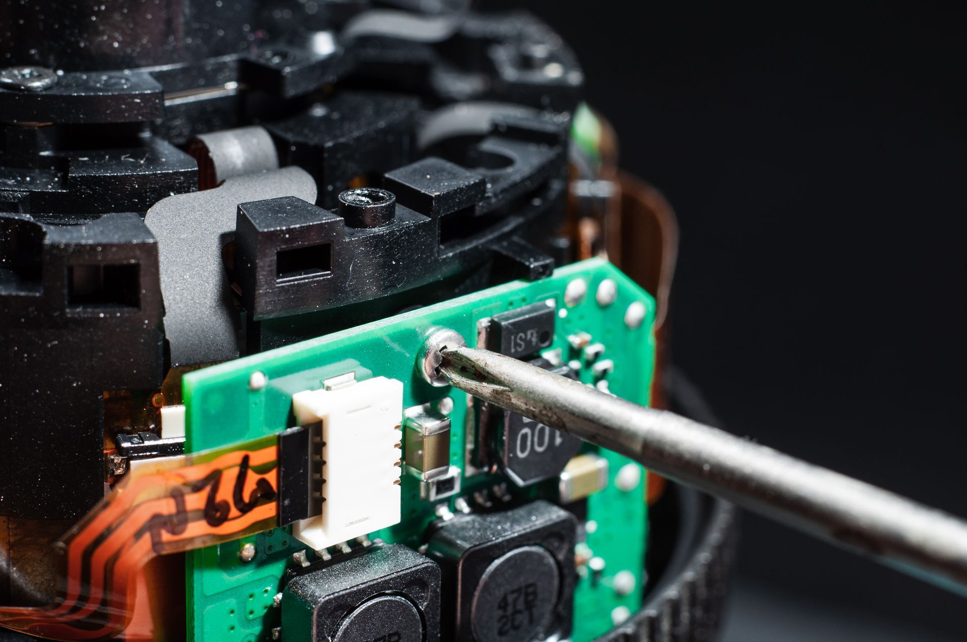 A metal screwdriver tip unscrews a small component on a green circuit board within disassembled electronic equipment.