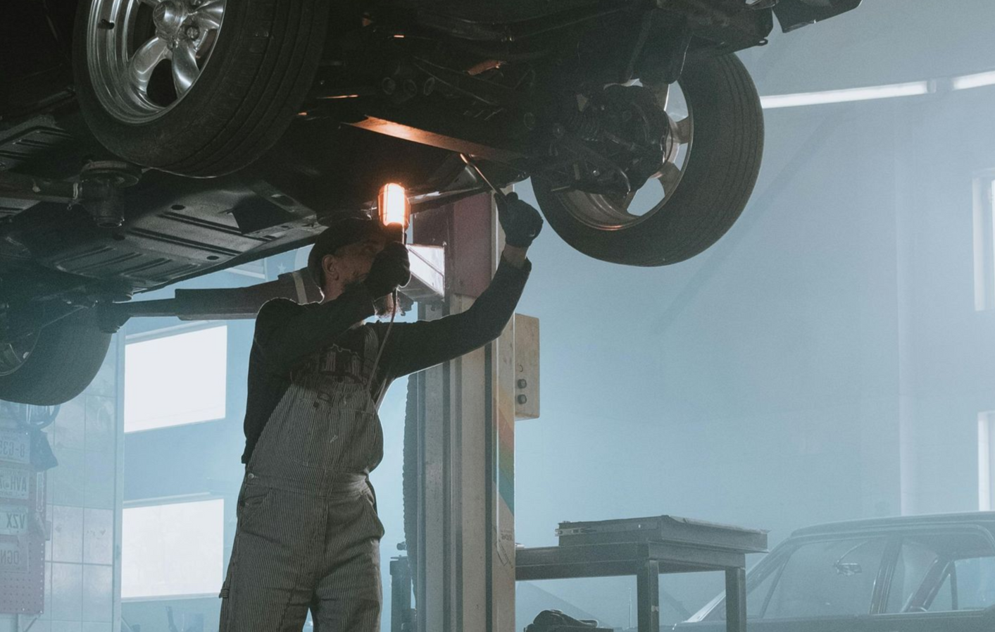 A mechanic works on the undercarriage of a car hoisted on a lift in a dimly lit, foggy workshop.