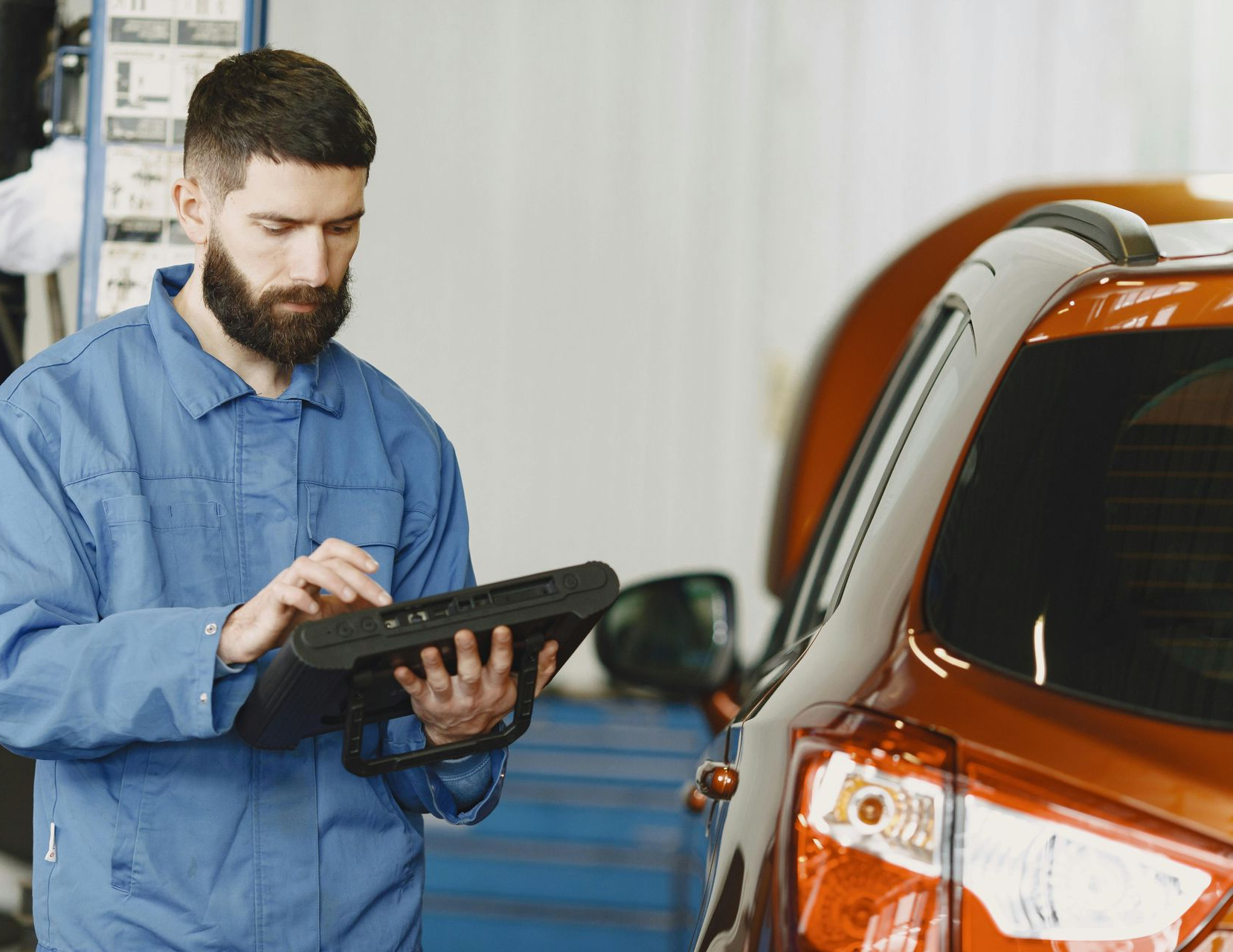 A mechanic in a blue uniform uses a diagnostic tablet while standing next to an orange car in a repair shop.