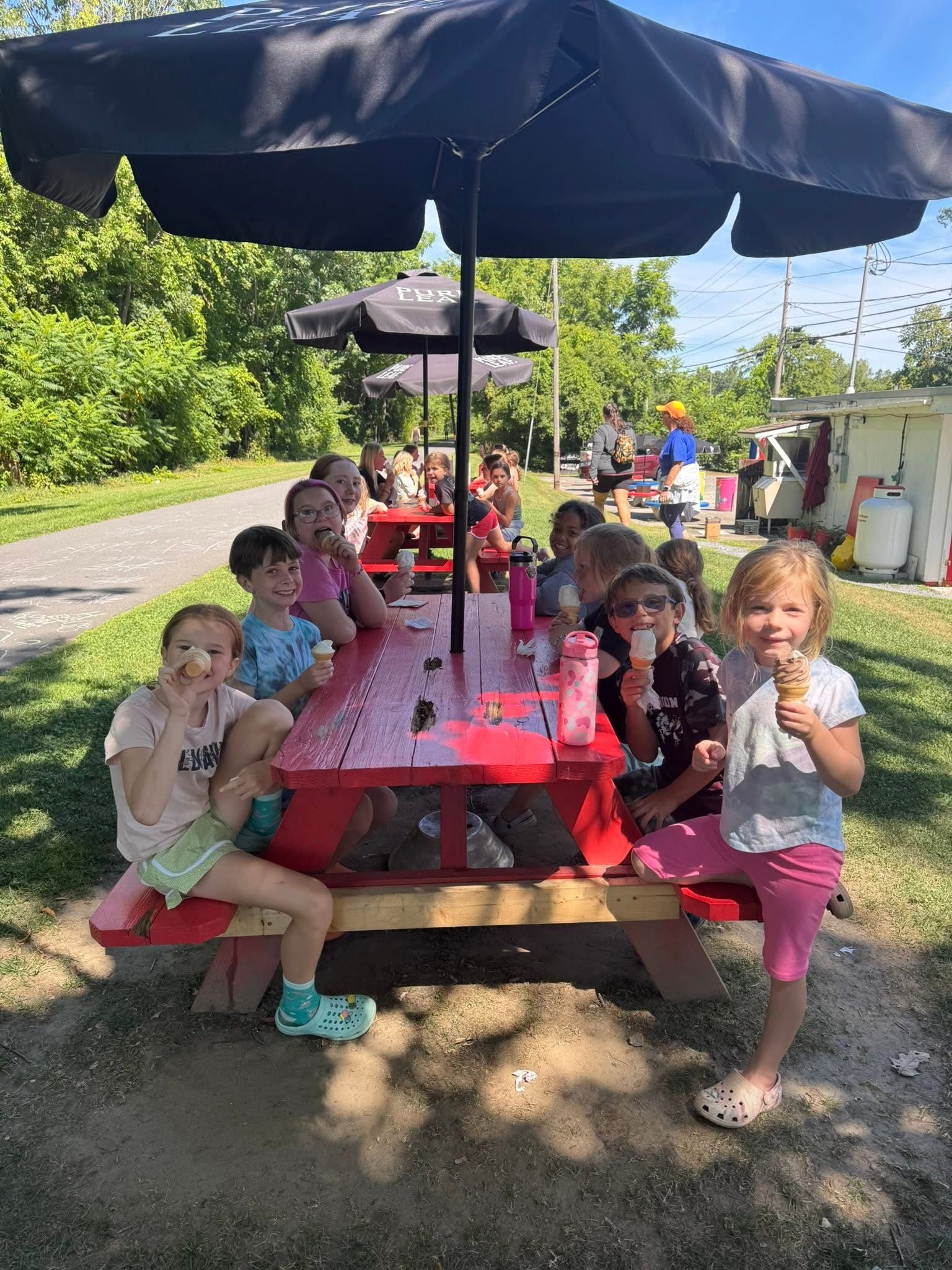 Children enjoy ice cream at a red picnic table under a black umbrella.