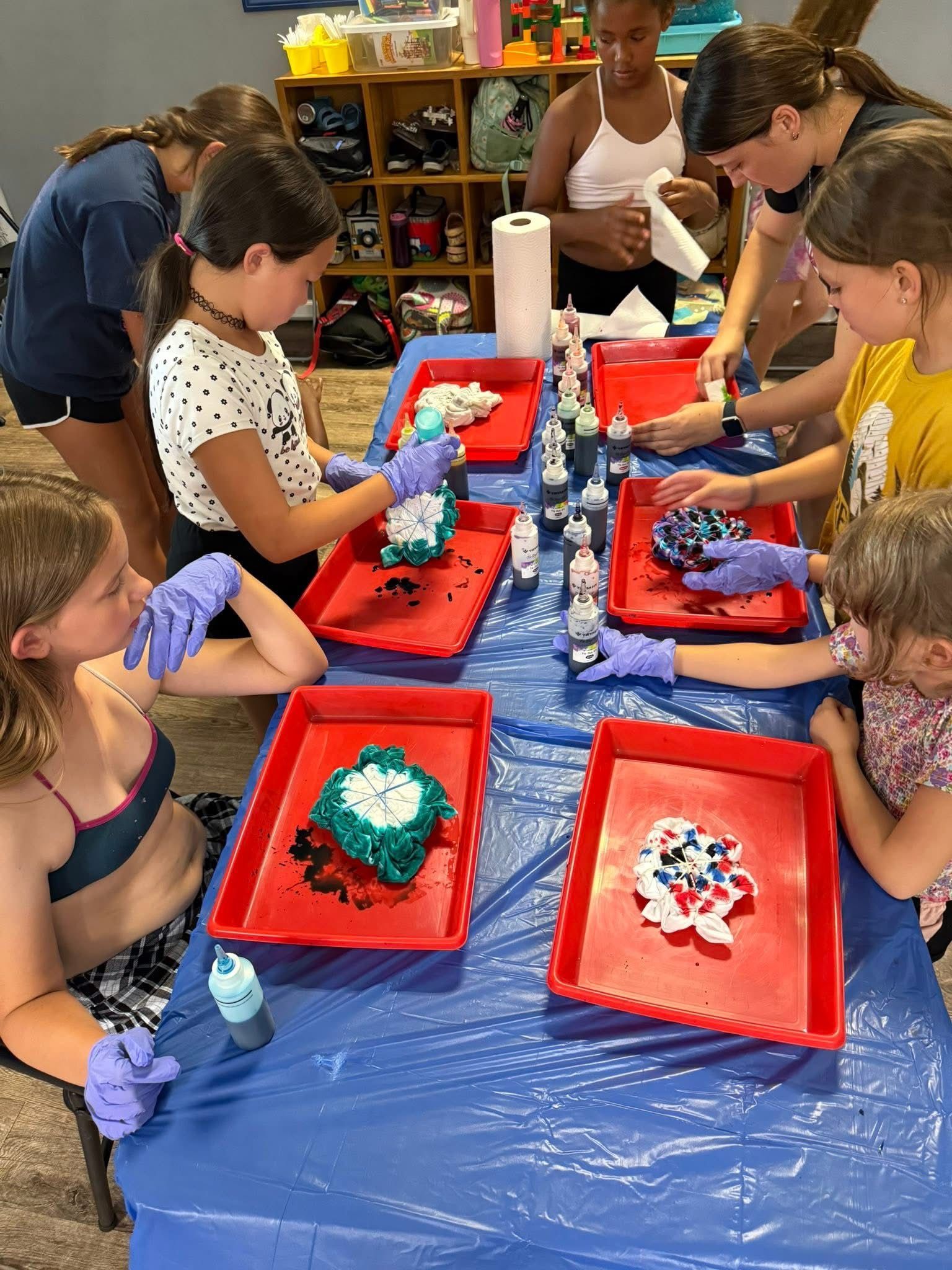 Children tie-dyeing fabric on a blue table. They use dye bottles and wear gloves. Indoors, with red trays.