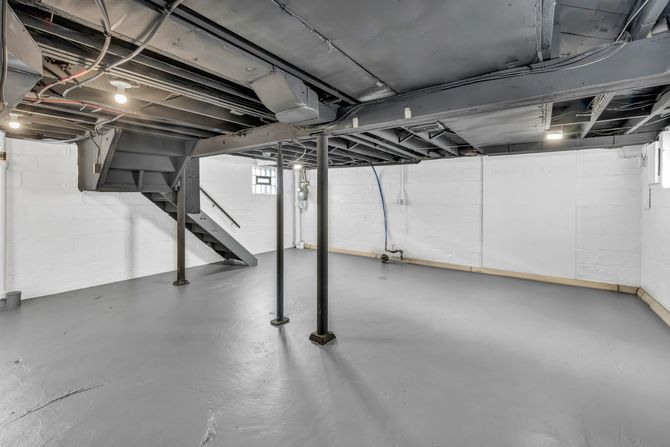 Empty basement with exposed ductwork, support posts, gray floor, and staircase along the left wall