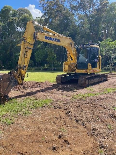 A yellow Komatsu excavator sits on a dirt construction site against a backdrop of trees and a bright, cloudy sky  — GPE Plumbers In Kendall, NSW