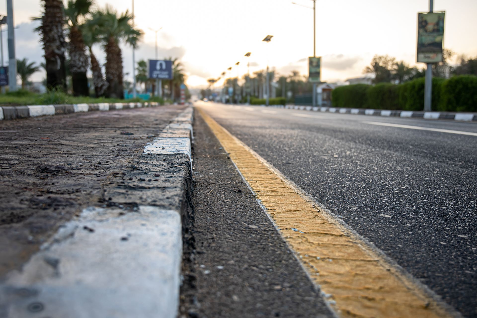 Low-angle view of a paved road with a yellow line and a black-and-white curb, alongside palm trees at sunset — GPE Plumbers In Kendall, NSW