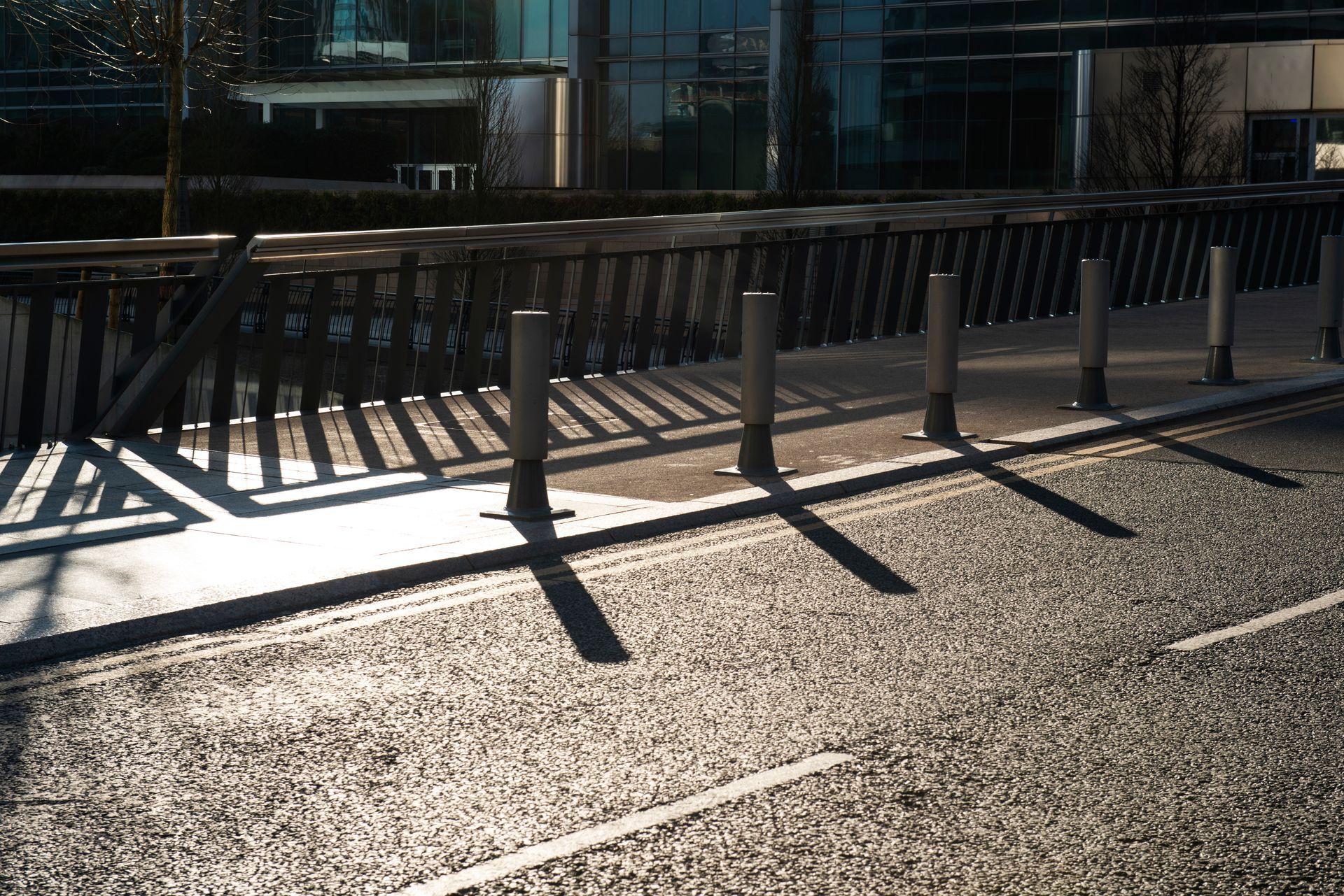 A row of metal bollards separates a sidewalk from an asphalt road, casting long shadows in the sunlight — GPE Plumbers In Kendall, NSW