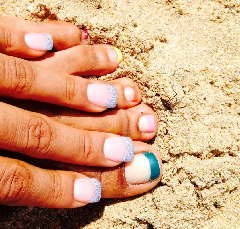 Close-up of toes on sandy ground, featuring manicured nails with pink, glittery, and teal French tip designs.