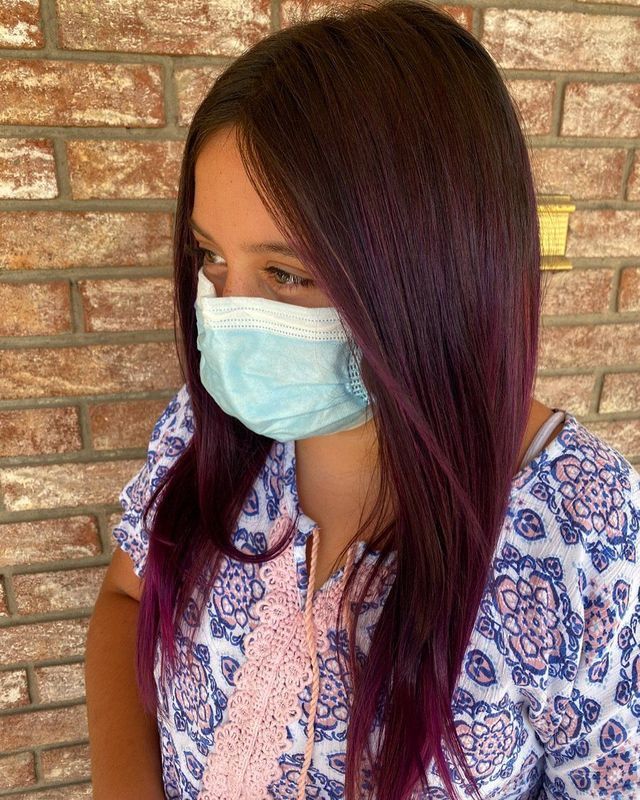 A person wearing a blue face mask with long, dark purple hair, standing in front of a light brown brick wall.