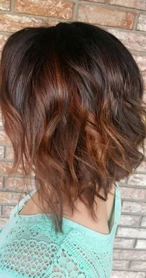 A woman with a dark brown, shoulder-length wavy bob hairstyle featuring copper-colored highlights against a brick wall.