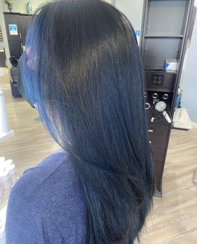 A back view of long, straight hair dyed a dark, midnight-blue shade in a hair salon setting.