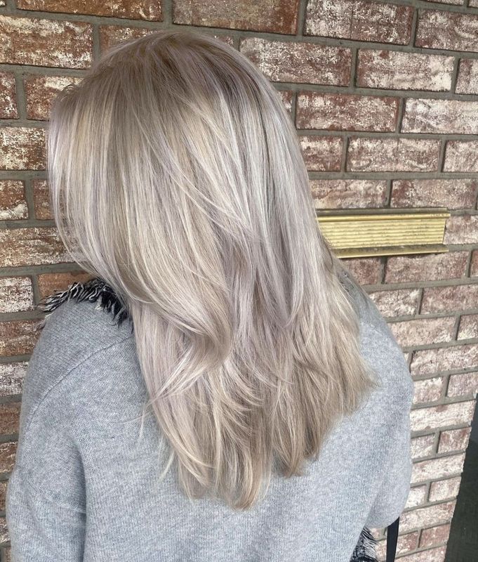 A person seen from behind, showcasing shoulder-length, layered, platinum-blonde hair against a brown brick wall.