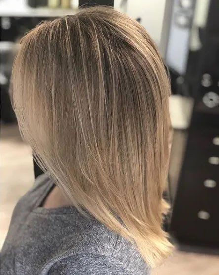 A person with layered, shoulder-length blonde hair, styled with a blowout and subtle highlights in a salon setting.