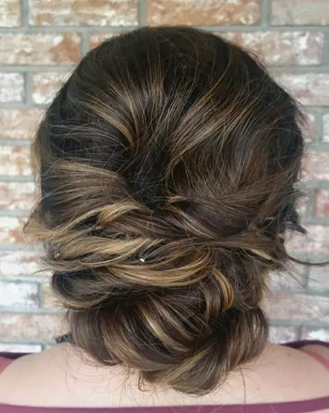 Back view of a person with brown, highlighted hair styled in a textured, loose low bun against a brick wall background.