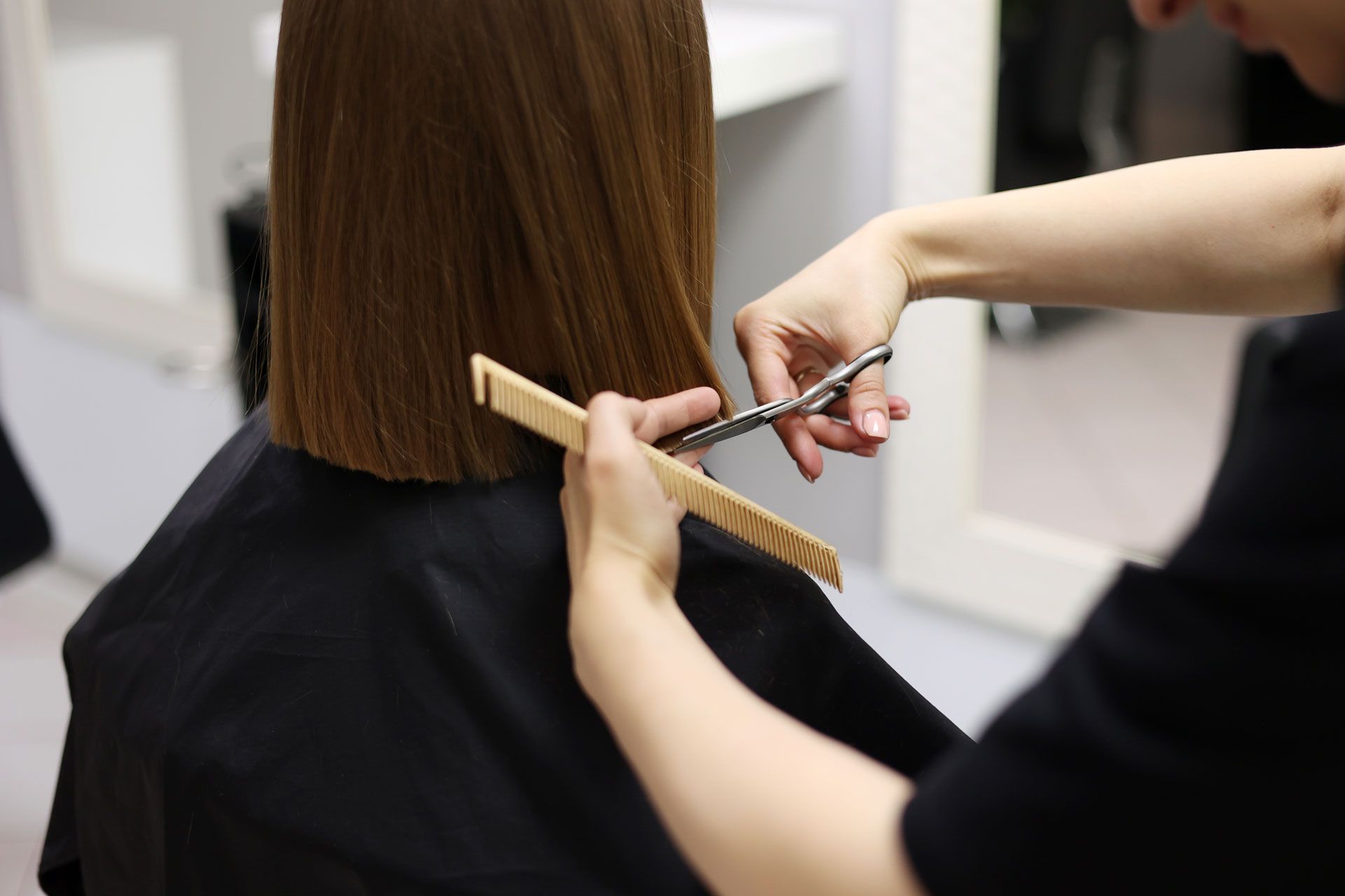 A stylist cuts a person's shoulder-length brown hair with scissors and a comb in a salon.