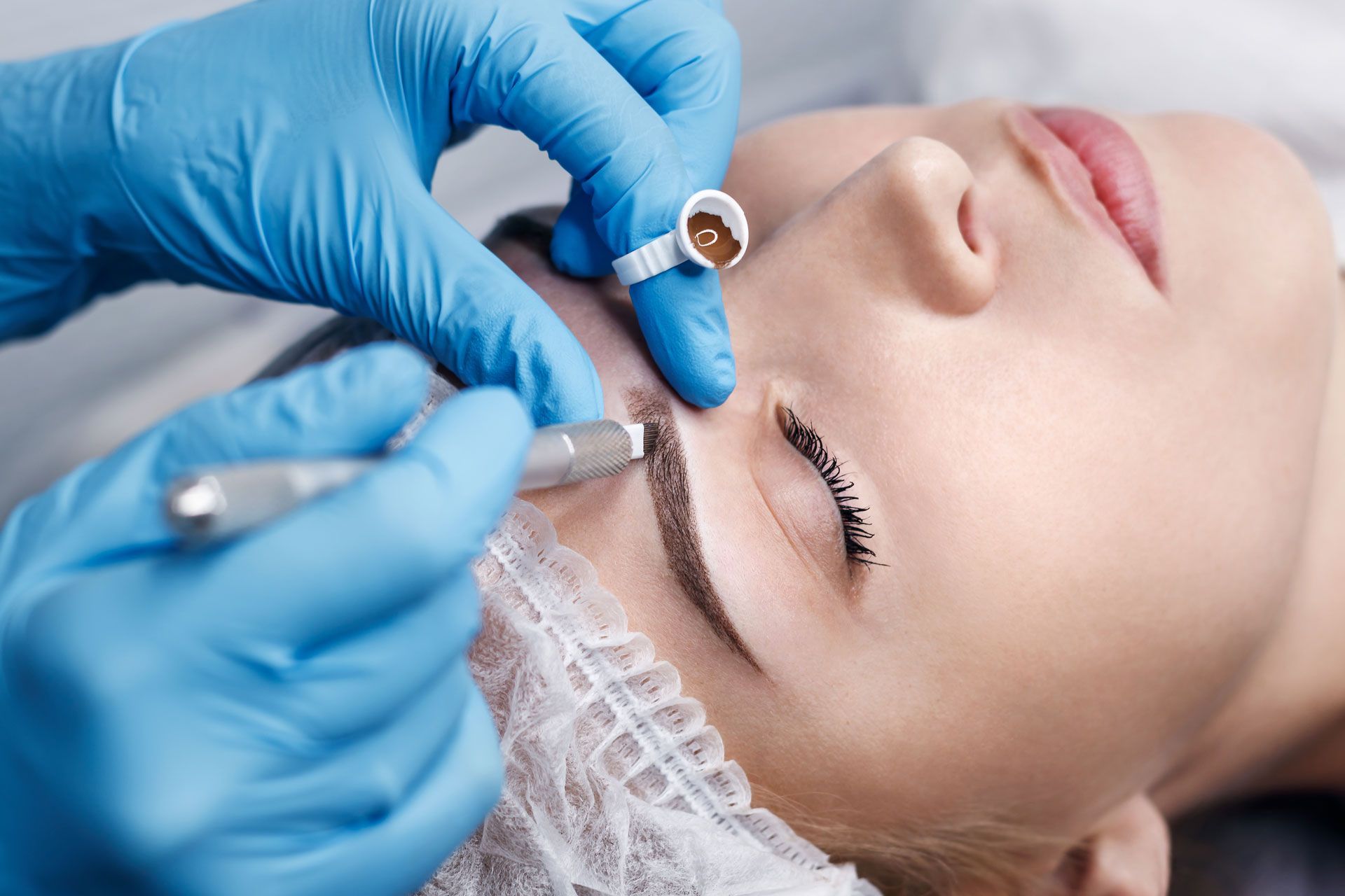 A technician in blue gloves performs microblading on a person's eyebrow using a specialized handheld tool.