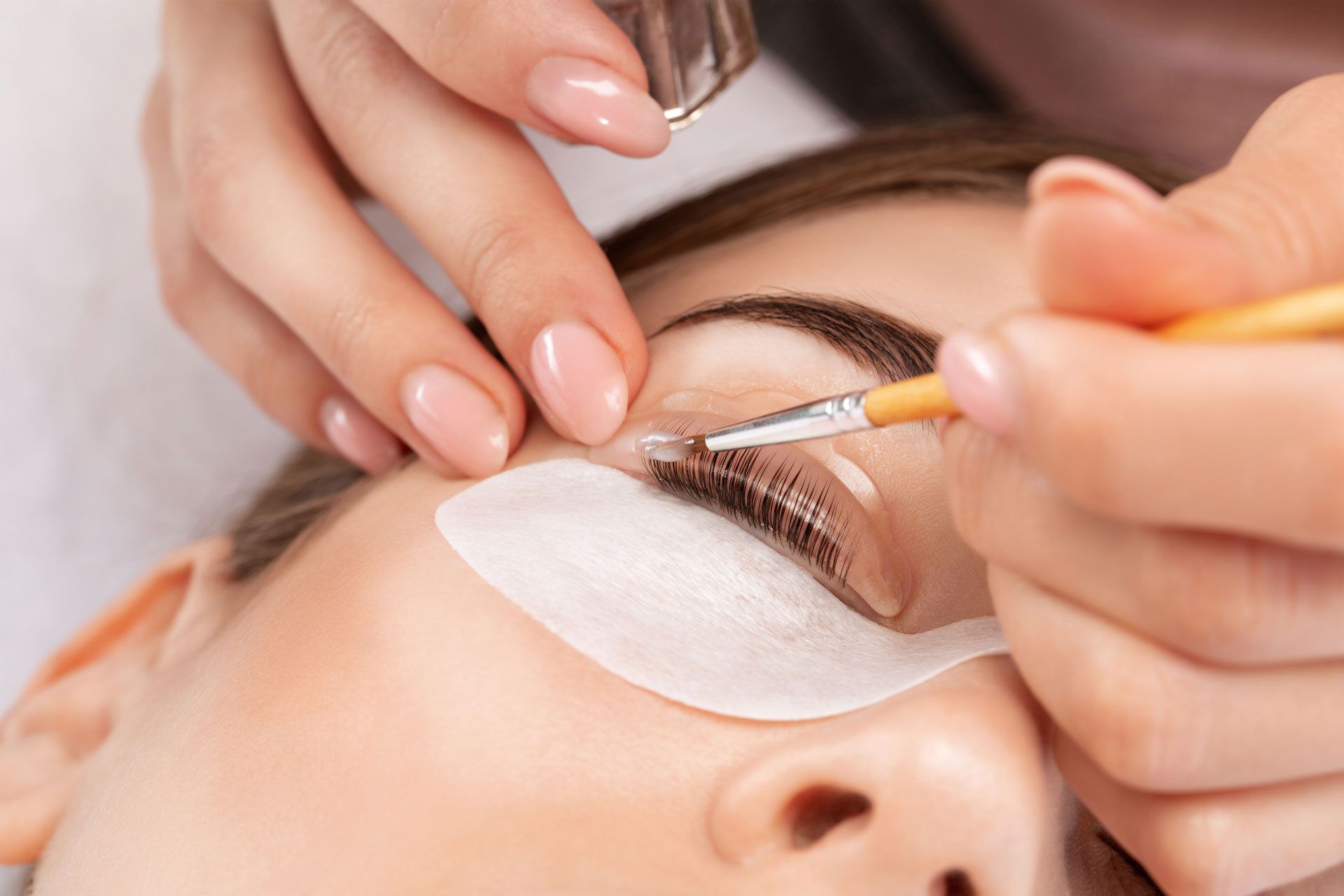 A technician uses a small brush to apply product to a person's eyelashes lifted onto a silicone pad.