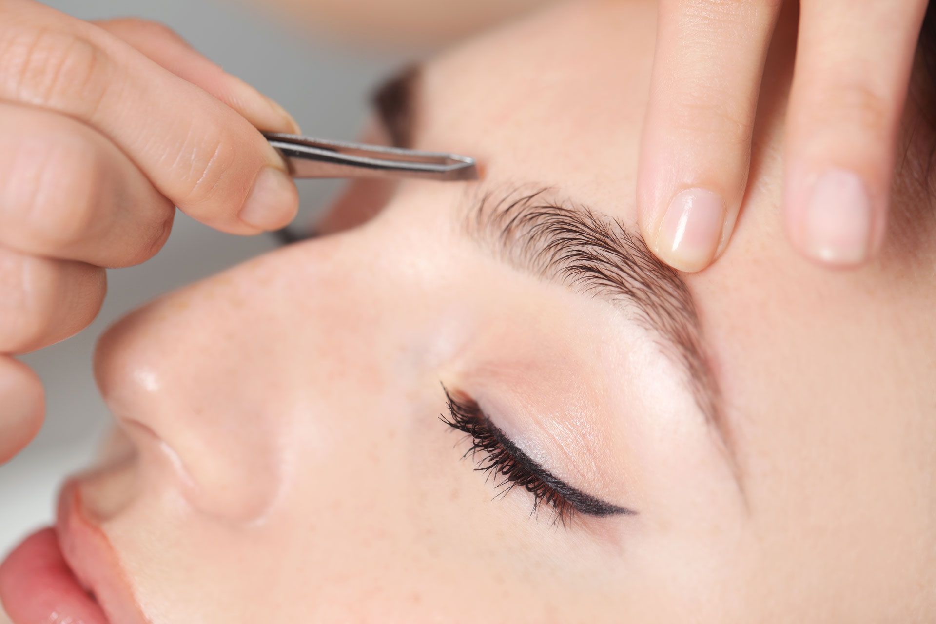 Close-up view of hands using tweezers to groom and shape an eyebrow.