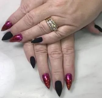 Hands with sharp, stiletto-shaped nails painted in alternating matte black and shimmering red-to-magenta glitter.