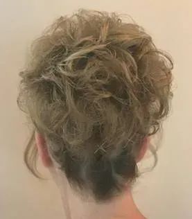 A back view of a person with blonde, curly, layered hair styled in an updo against a plain light-colored background.