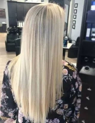 A person with long, straight, platinum blonde hair wearing a black floral top, seen from behind in a hair salon.