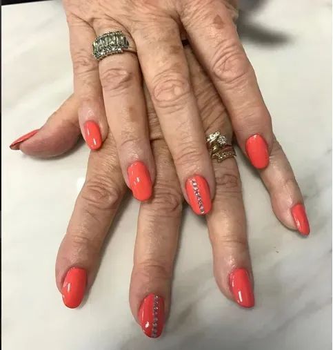 Hands with bright coral manicured nails, some accented with vertical lines of small rhinestones, wearing two gold rings.