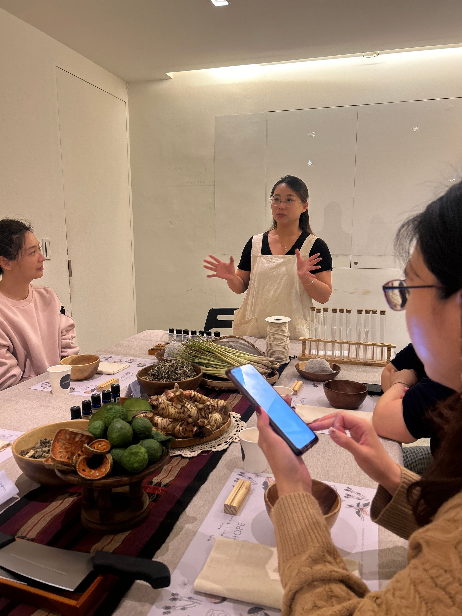 身穿白色洋裝的女子，手持木碗，在戶外綠樹成蔭的地方，聞著一個小瓶子的味道。